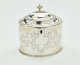 A silver tea caddy with a lid and sculptural finial. There are swirling engravings along the body and lid.