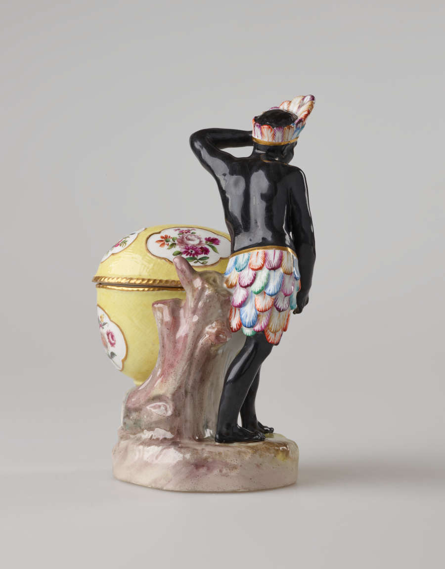 A sculptural figure with black skin, a colorful headdress and skirt. This figure is standing next to a bright yellow covered sugar basket with floral decorations.