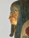 Side profile of a gilded coffin face with blue-outlined eyes, a sculpted false beard, and an ornate headdress with gold detailing and visible signs of age and wear.
