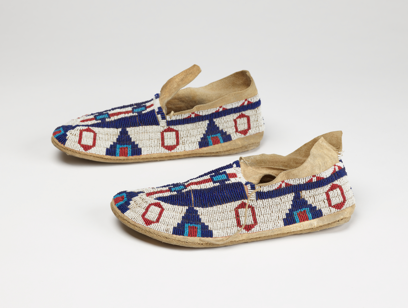 Pair of fabric shoes side-by-side and in profile, made from beaded fabric with geometric patterns in red, deep and light blues. The moccasins’ mouths are open and have two laces.