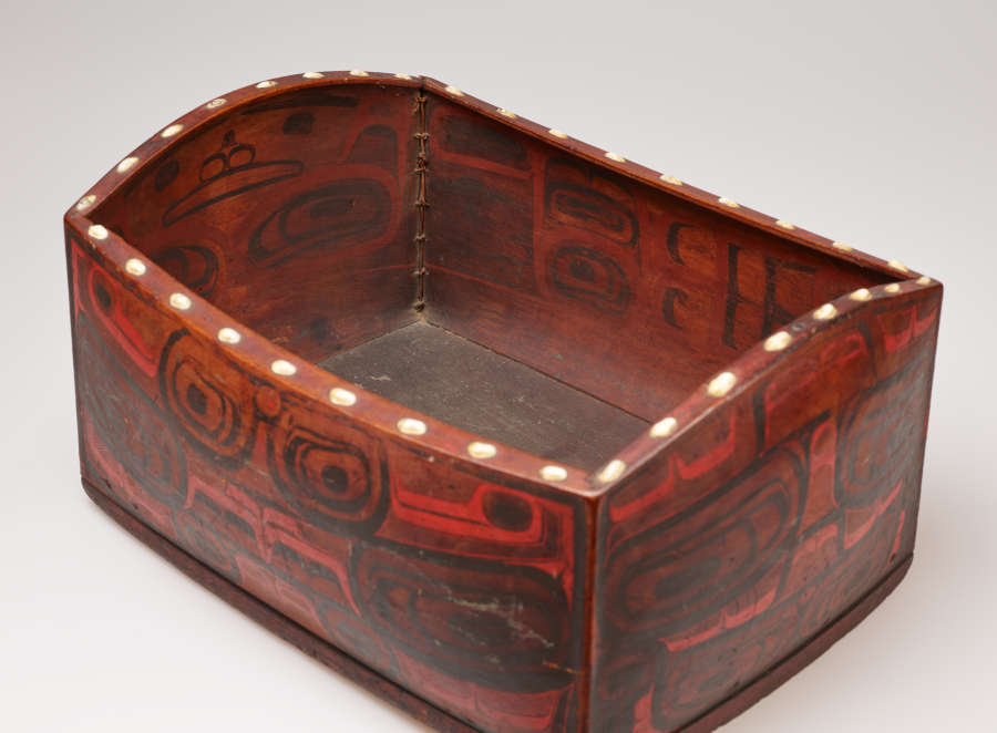 Quarter-view of a bulging rectangular wooden box with faded, red and black, graphic illustrations, some mimicking faces. The box's rim undulates and is decorated with a line of white dots.