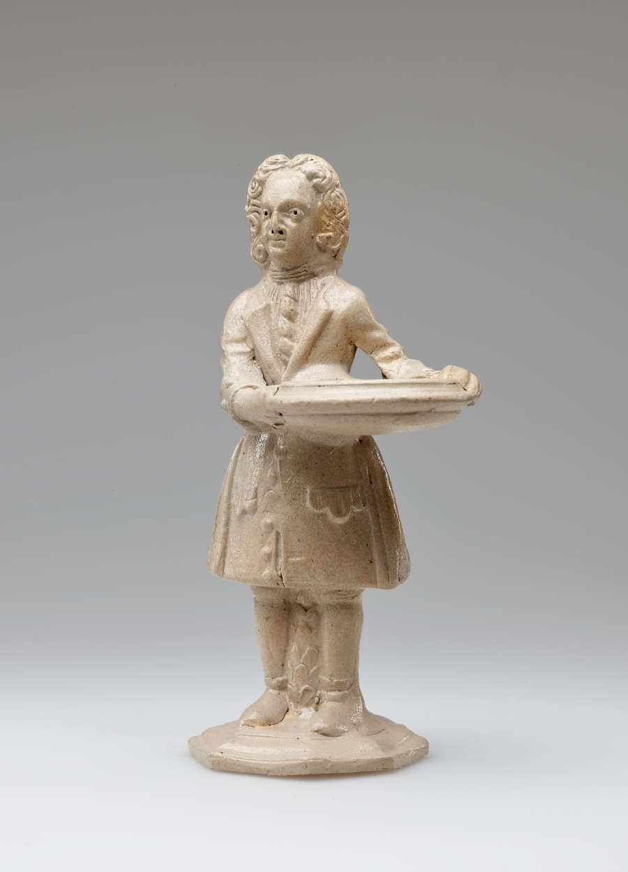 A cream colored sculptural figure holding a vessel, dressed in historical clothing with tightly curled hair.
