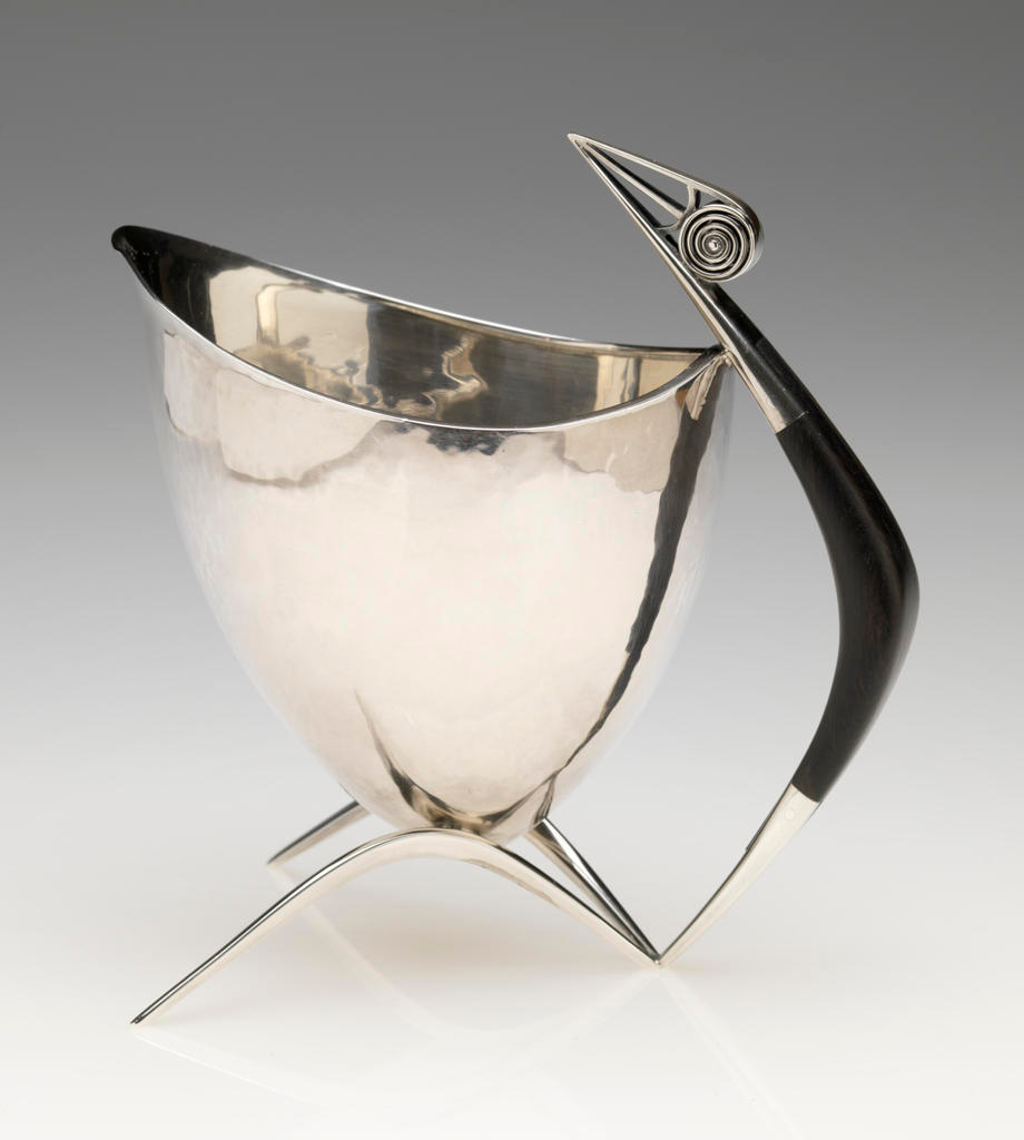 Sleek silver vessel with a curved body atop three pointed legs, featuring a black handle with a spiral detail and sharp ends which stretch above the vessel's top.