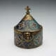 Copper box with cylindrical bottom and conical lid with gold and blue enameled geometric and floral patterns, topped with a carved cross.