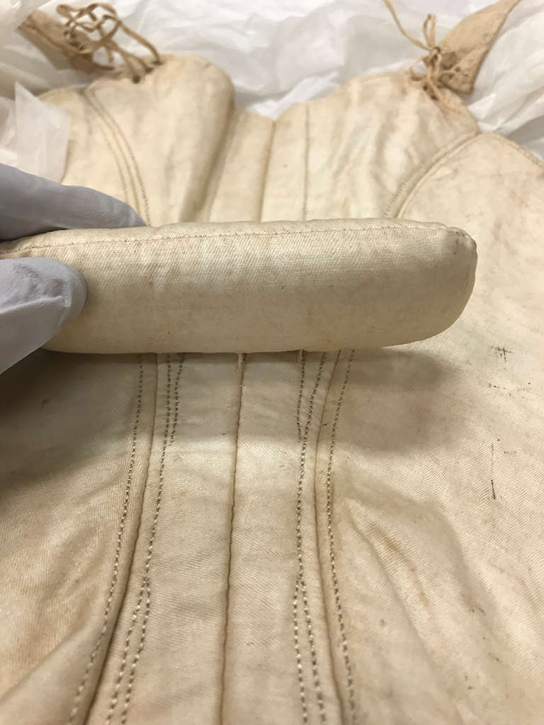 A close-up of a lifted padded fabric roll attached to a cream-colored fabric corset with curving boning and visible stitching.