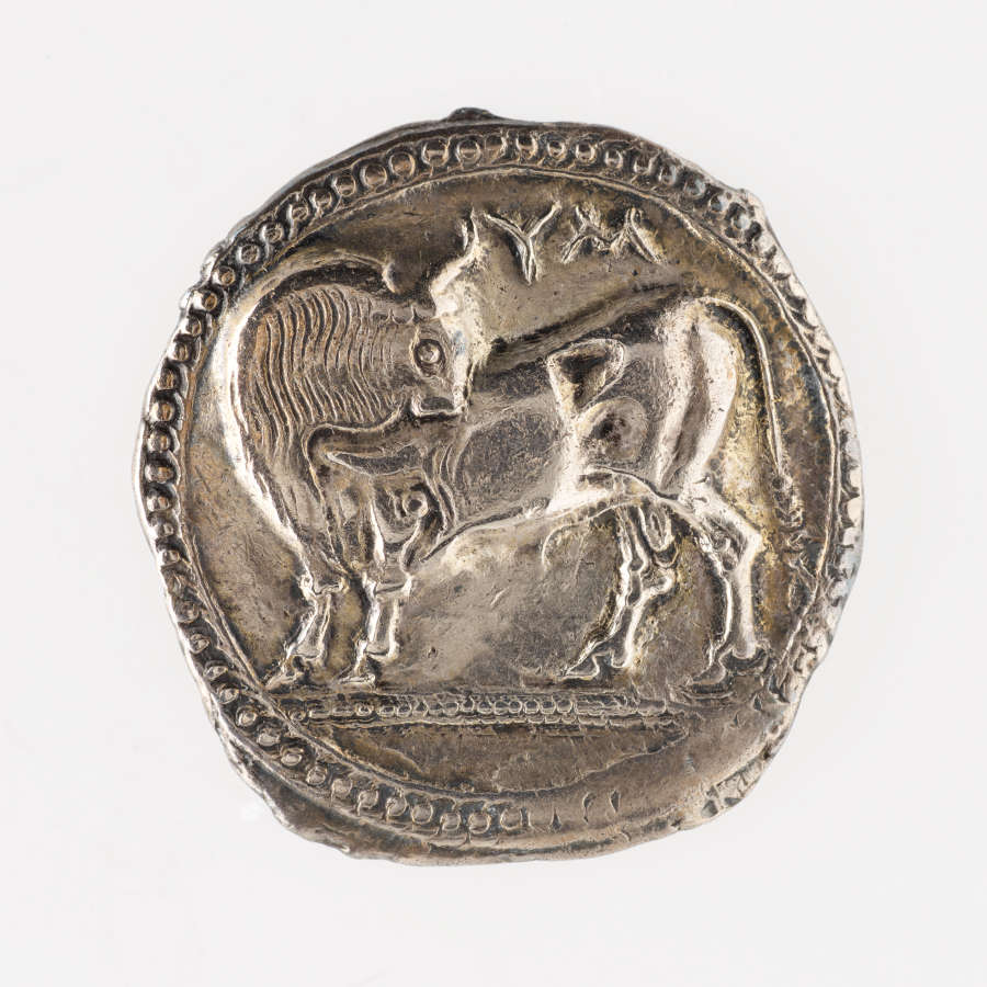 Silver coin with irregular borders. Embossed on one side is a four-legged creature turning its head. The border consists of small raised half-spheres.