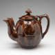 A mottled brown teapot with an image of a pineapple in the center. Two spouts are on the left side, and a rounded handle with a flat top faces right.