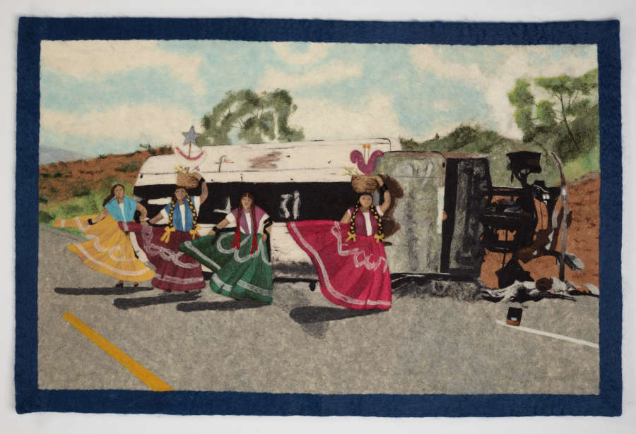 Colorfully dressed figures dancing in traditional Mexican clothing. They are positioned in front of a destroyed bus on the side of a road, against natural scenery with a clear sky.  
