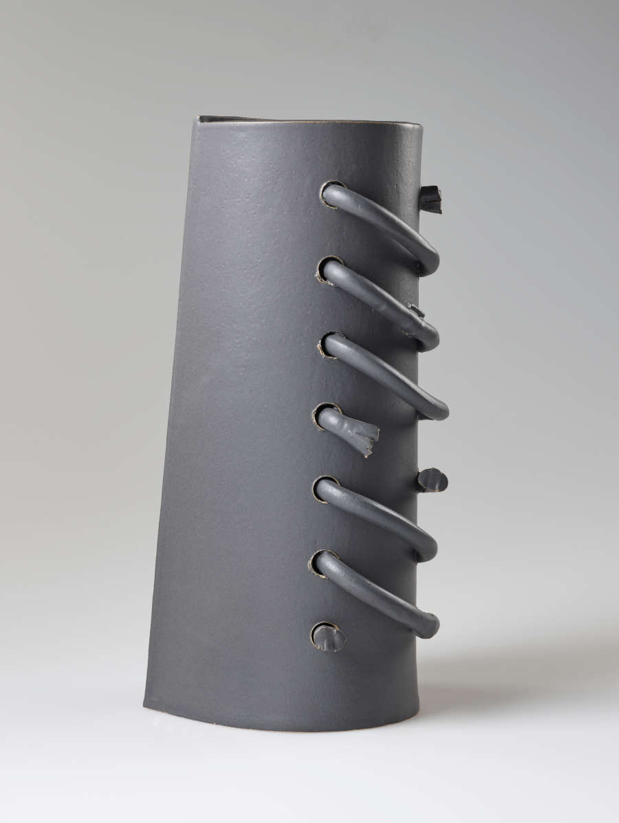 Diagonal back view of a cylindrical gray sculpture featuring a row of helical protruding loops threaded through the ceramic surface. Some of the loops are broken.