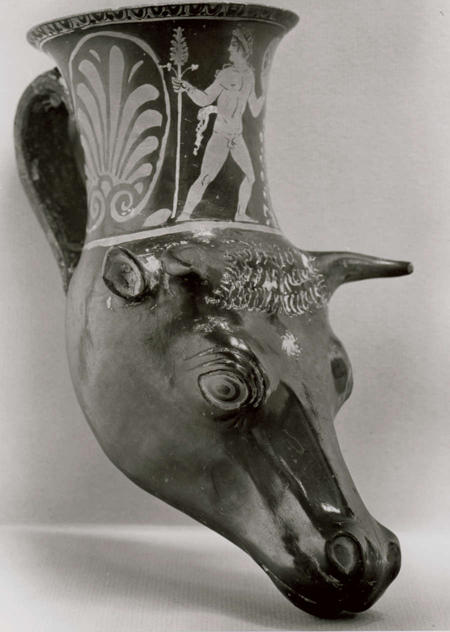 Monochrome photo of a cup with the base in the shape of a cow’s head, its top extending into the fluted body of the cup, which is painted with illustrations of men next to large floral motifs. 