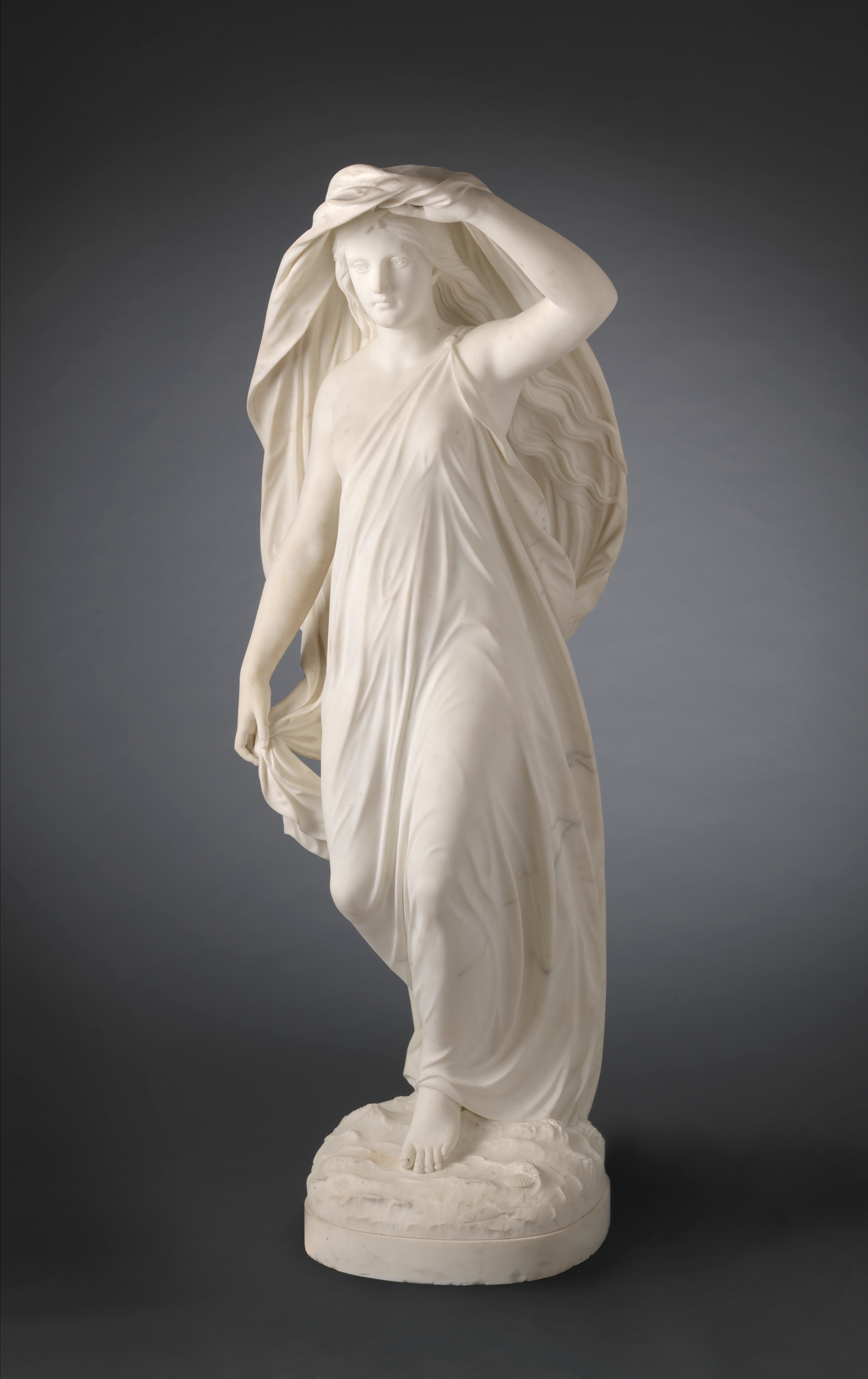 A marble statue of a figure draped in flowing fabric, lifting a veil over the head and stepping forward.