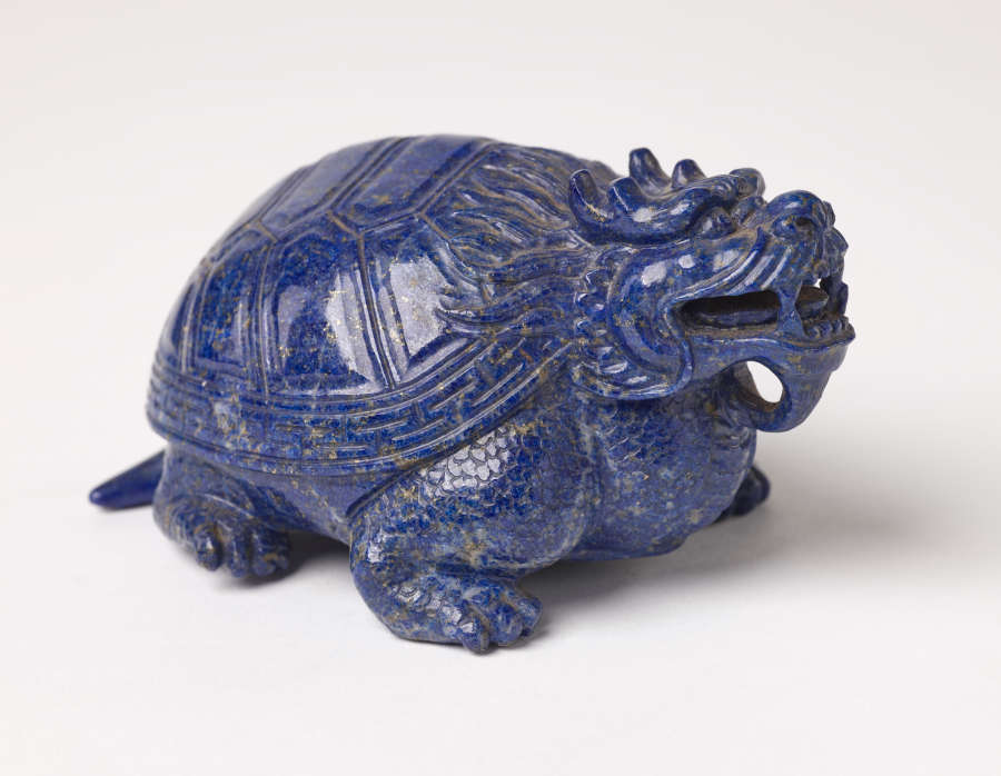 A quarter view of a blue sculpture of a horned tortoise, whose surface is covered with carved geometric patterns.