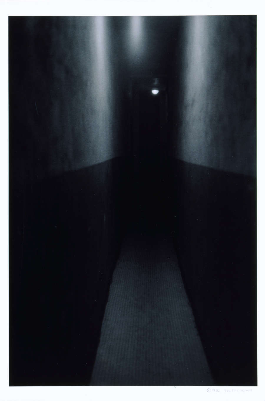 An eerie, dimly lit corridor with one unsettling white light bulb at the end and tall walls stretching back into darkness. 