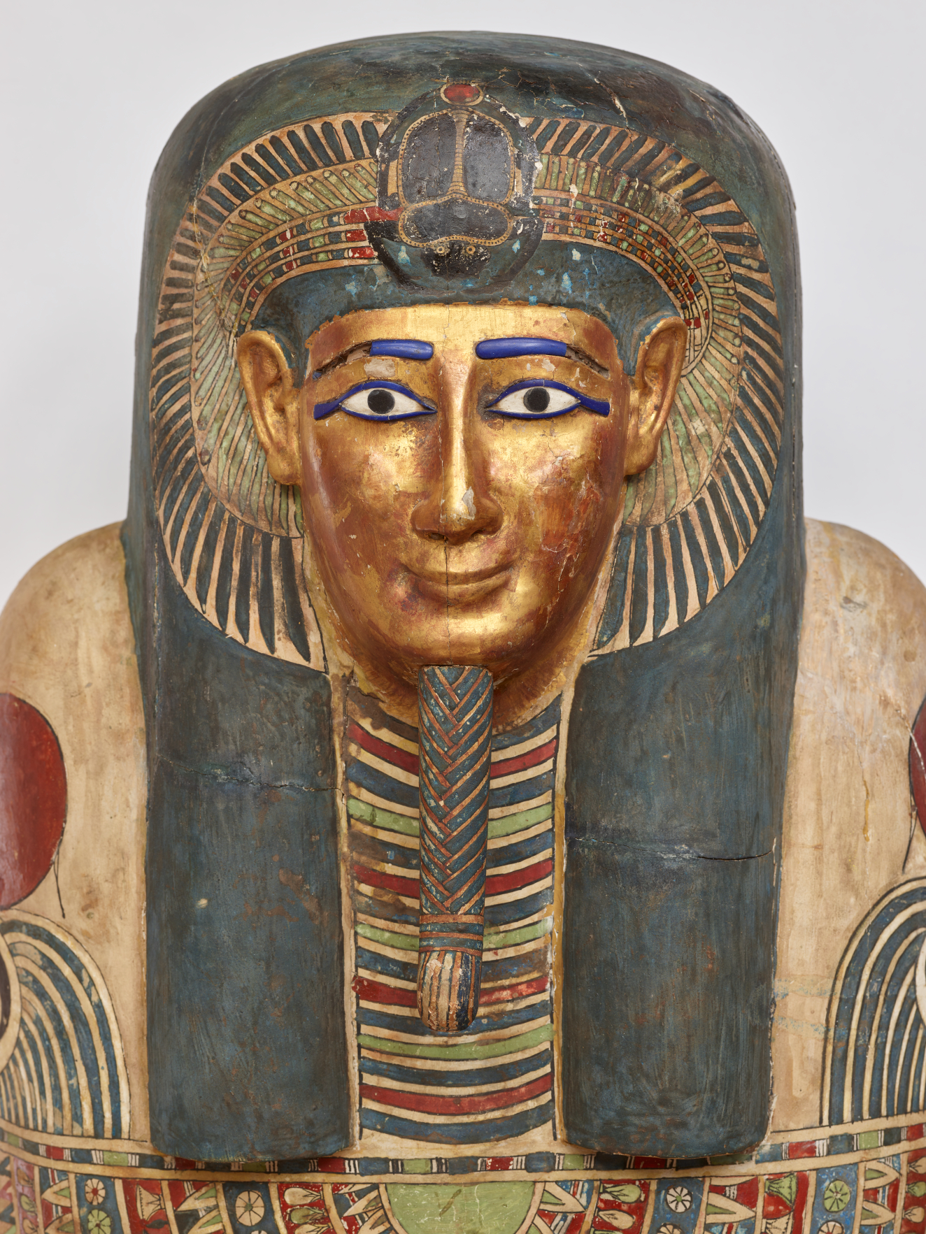 A gilded face with blue-outlined eyes on a coffin lid, framed by a blue black headdress with golden details and a red, green, and blue painted collar.