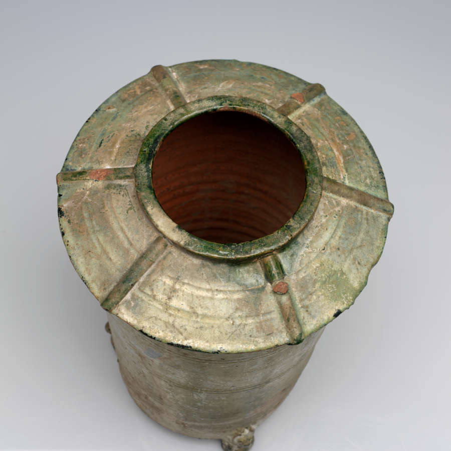 Top-view of a worn green-gray terracotta cylindrical urn showing its narrow circular mouth flaring into its wide and shallow saucer-like neck. 6 ridges radially branch off from the mouth's edge.