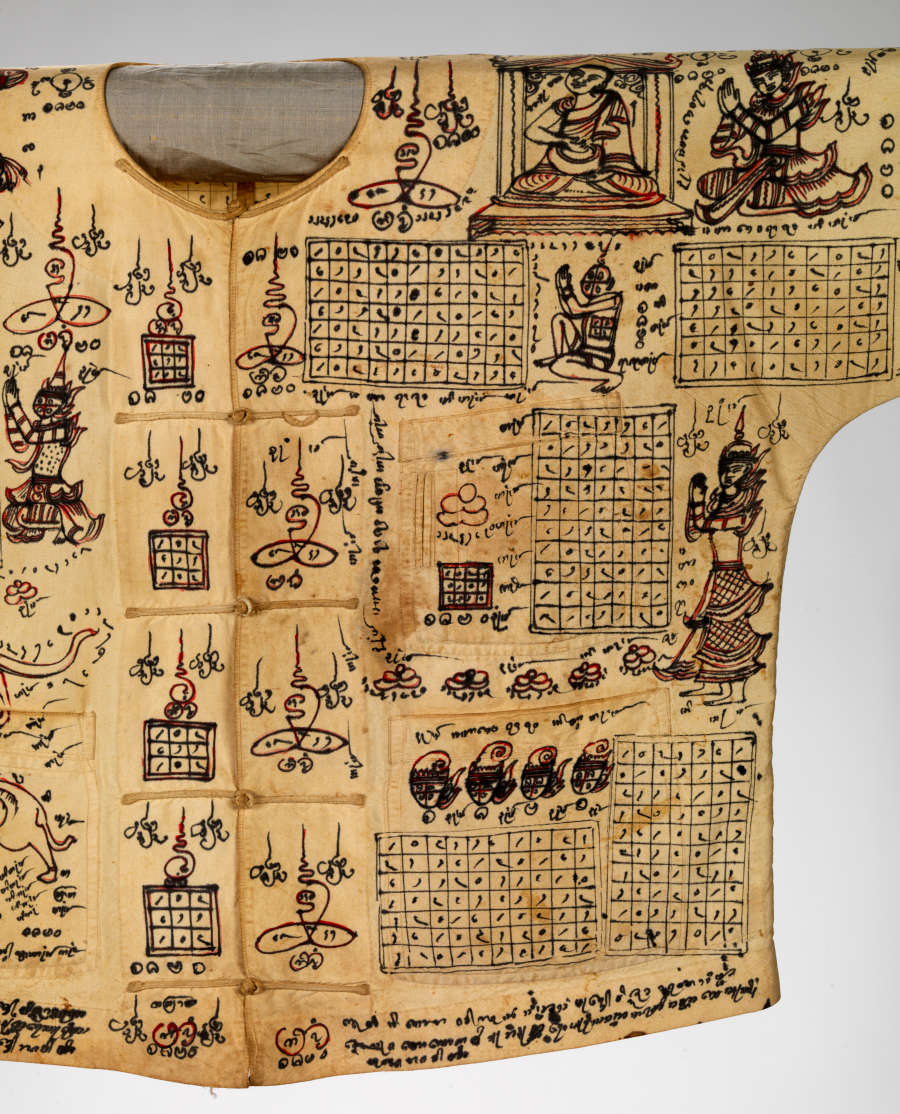 Detail-view of the yellowed shirt’s front, populated with illustrations of mythical characters, grids and symbolic writing in red and black. Its placket is secured by ties and has pockets.