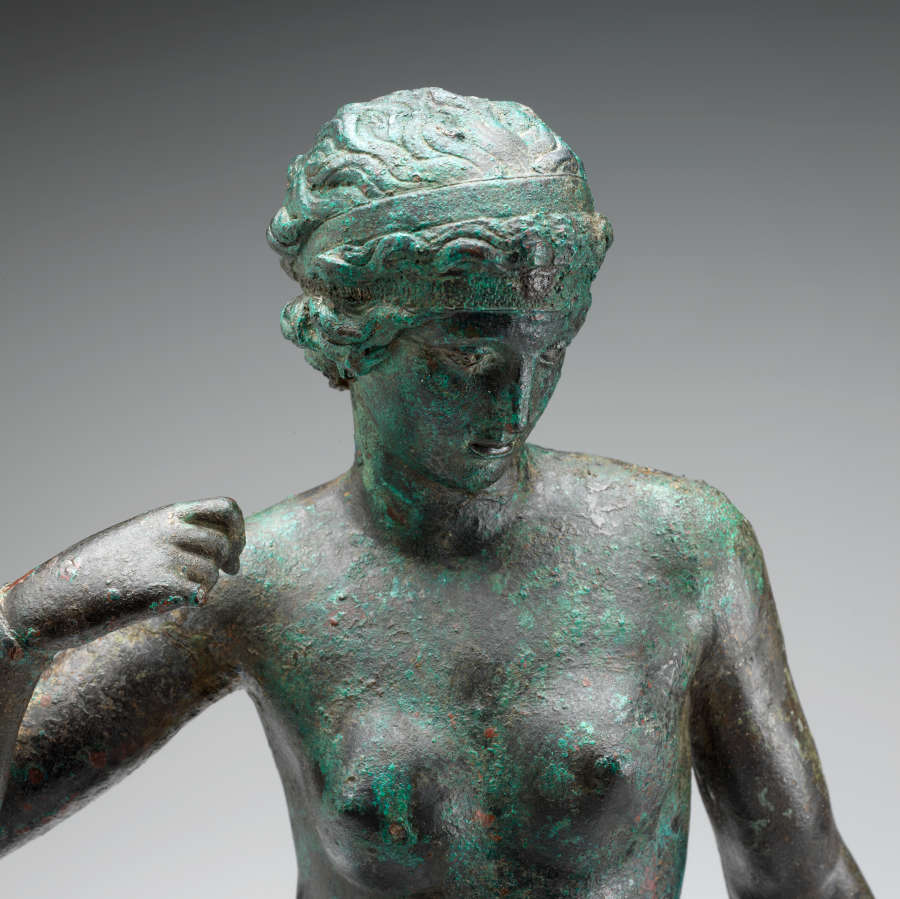 Detail of a bronze statue of a nude woman. Visible are the details of her face, hair and fist curled towards her shoulder as she looks slightly downwards.