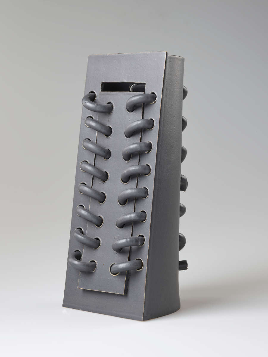 Diagonal front view of a rectangular gray ceramic sculpture featuring a row of horizontal loops threaded and protruding from the surface. The back features forms organized in a similar manner.