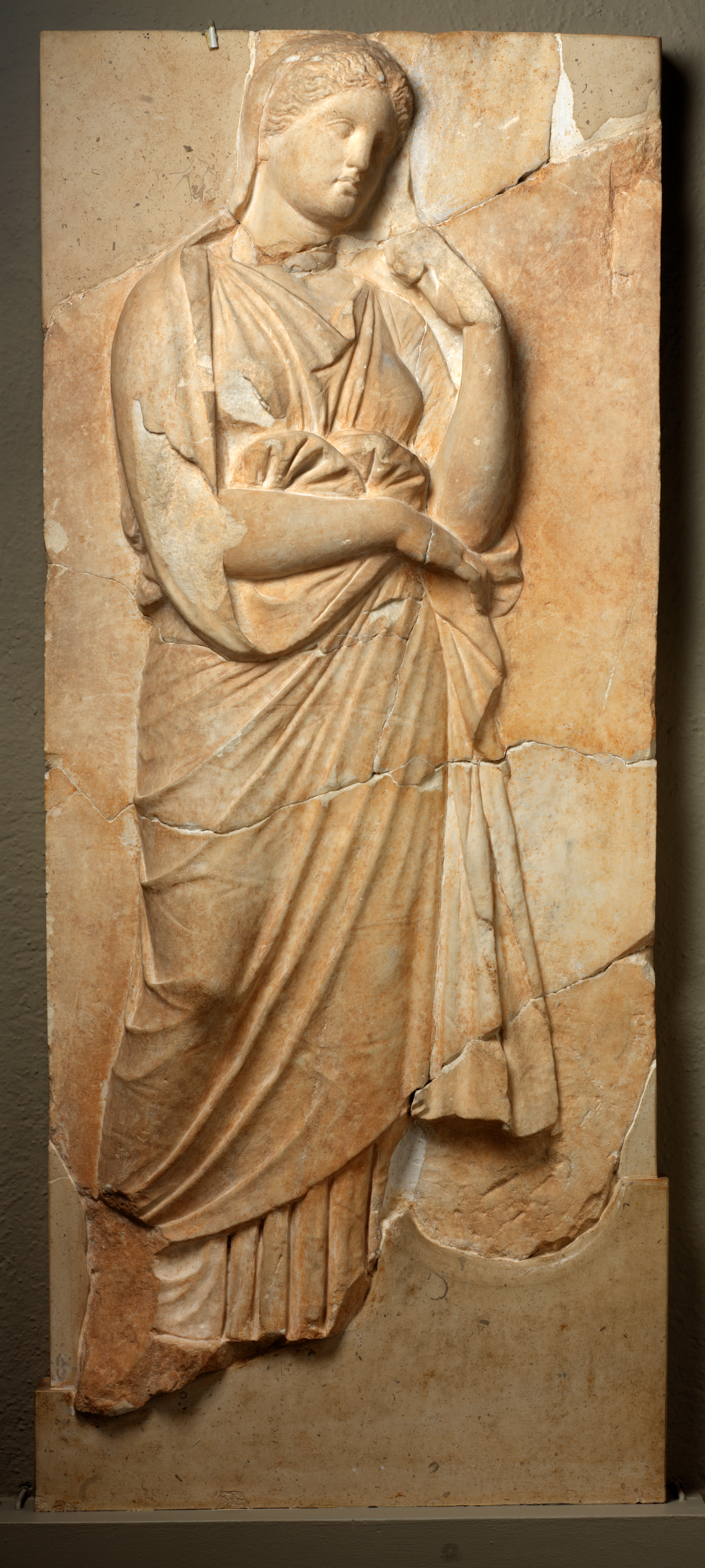 Beige stone carving of a figure in long, draped robes in a contemplative pose with head slightly tilted and one arm propped near her face, framed by cracked stone sections