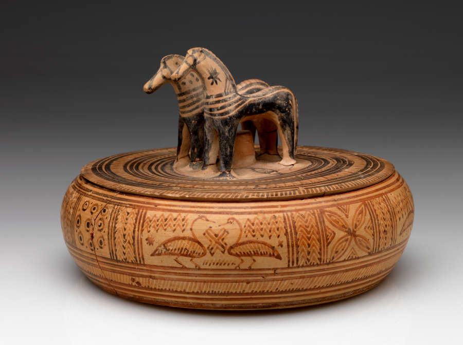 Side-view of a light-brown, short, wide, and round lidded box with a knob shaped as a pair of horses, decorated with patterns and natural imagery in black and red.
