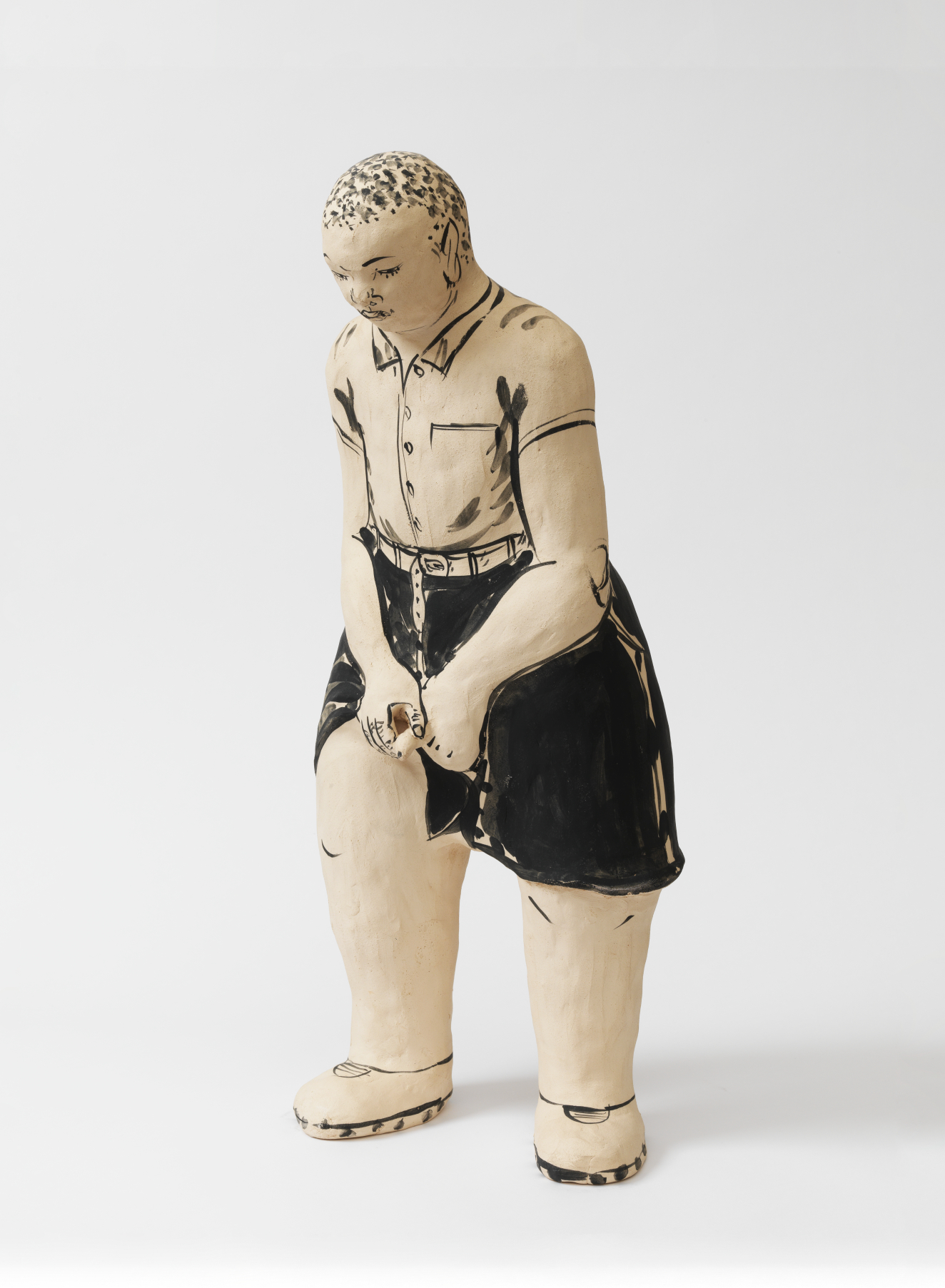 Stylized ceramic figure of a young boy leaning forward in a buttoned shirt tucked into dark shorts with his hands clasped in front of his pants, head tilted downward.