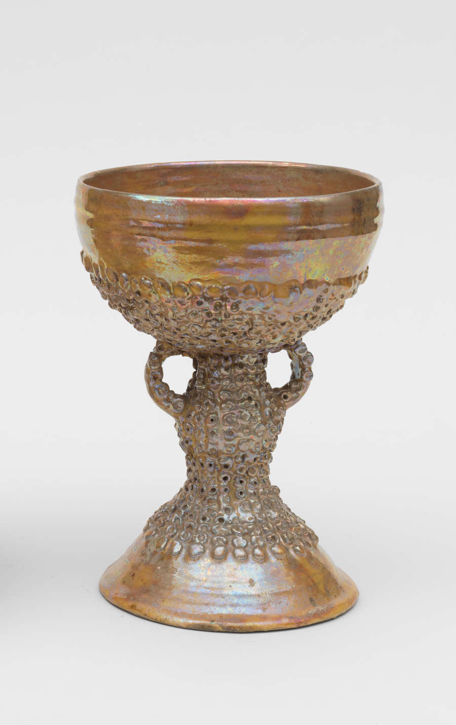 Golden metallic cup with a glossy, reflective surface, featuring a wide bowl and two small, looped handles joined to a thick textured stem that flares out into a circular base.
