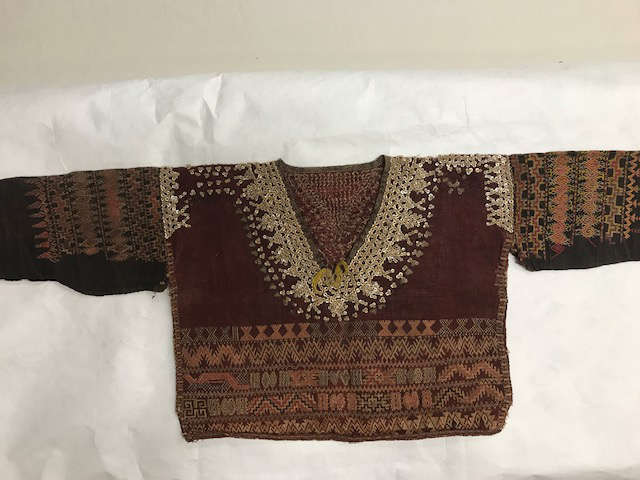 Dark red garment on a white background with intricate sequin embroidery around the neckline. Along the bottom and shoulders are dense stripes of geometric patterning in waves and triangle motifs. 