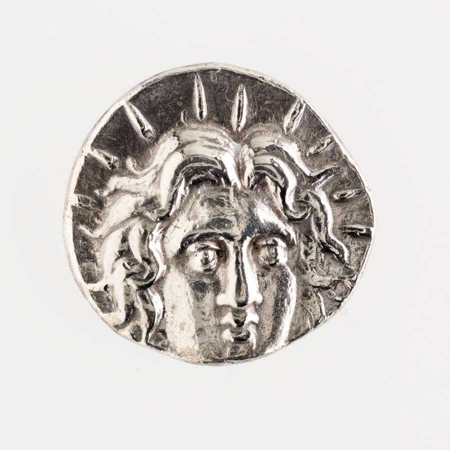 Silver coin with irregular edges. Embossed on its surface is a portrait of a person with wavy hair, lines emanating out from their profile.