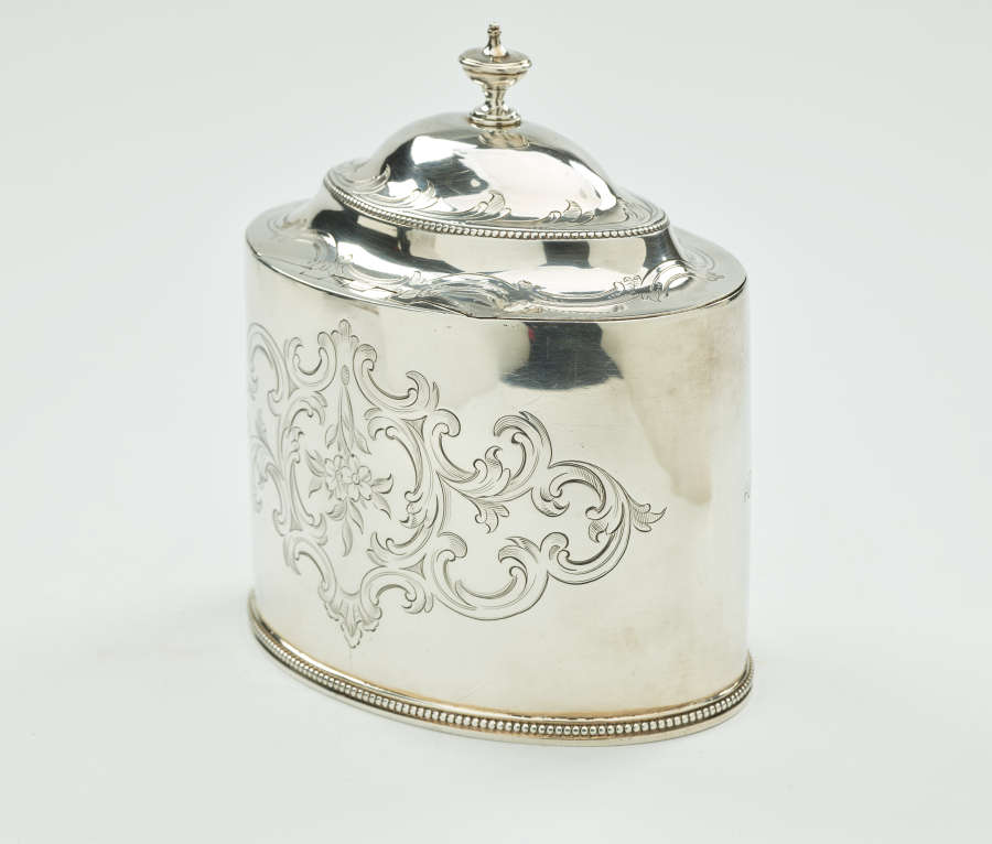 A silver tea caddy with a lid and sculptural finial. There are swirling engravings along the body and lid.