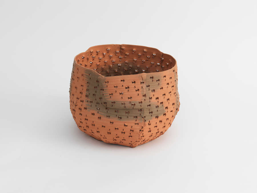 A small terracotta bowl with an uneven rim and a matte finish, the surface blending reddish and gray hues and punctured with tiny holes that form an irregular grid.