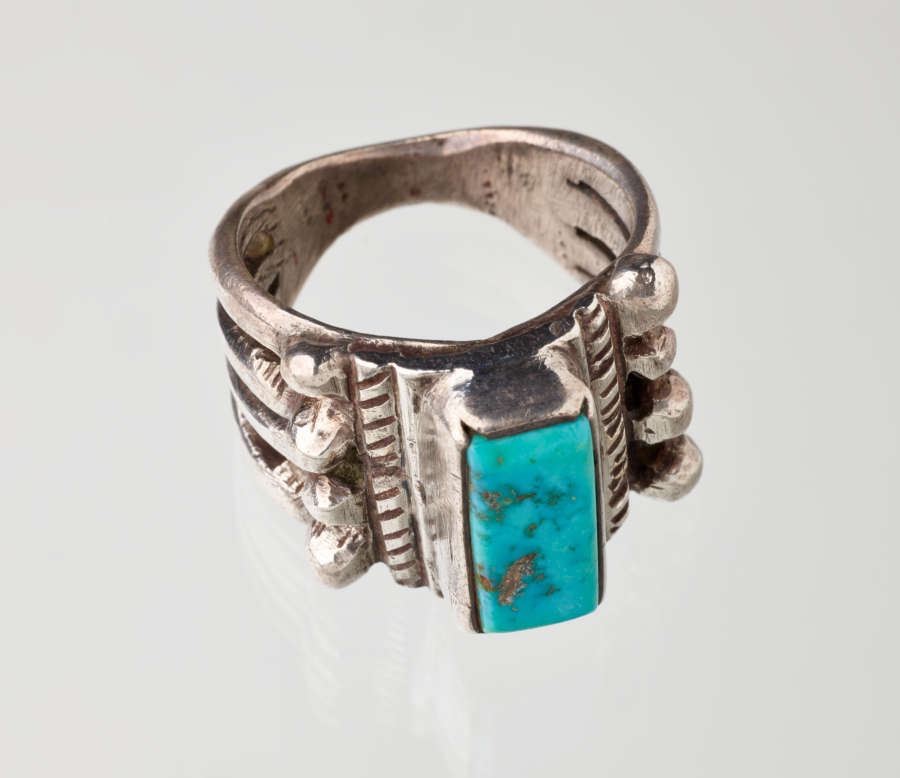 Three-quarters view of a silver metal ring with a rectangular turquoise stone setting embellished with nature-inspired and rope-like decorative elements. The band narrows towards its back.