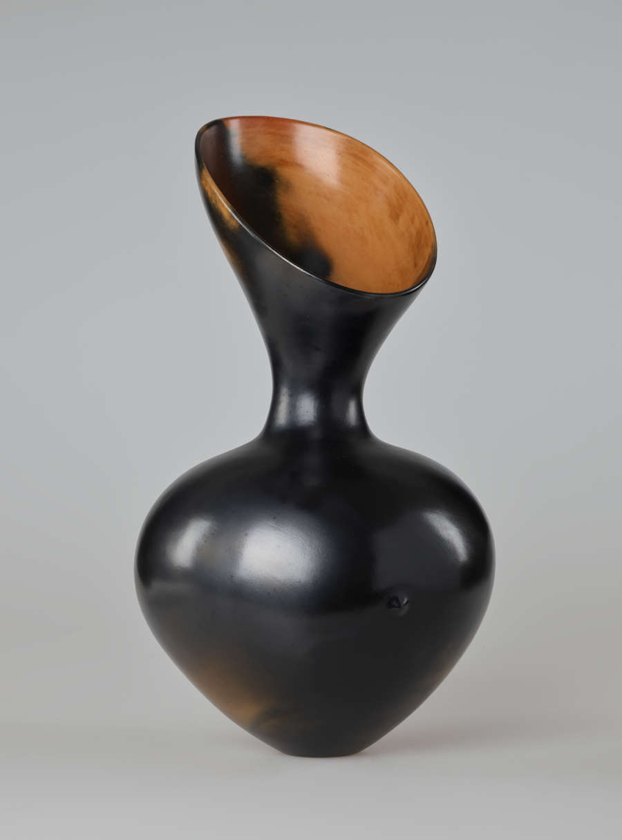 An asymmetrical, hourglass-shaped black vessel with a reflective finish. Its wide opening slopes down to the right, exposing a bright terracotta interior.