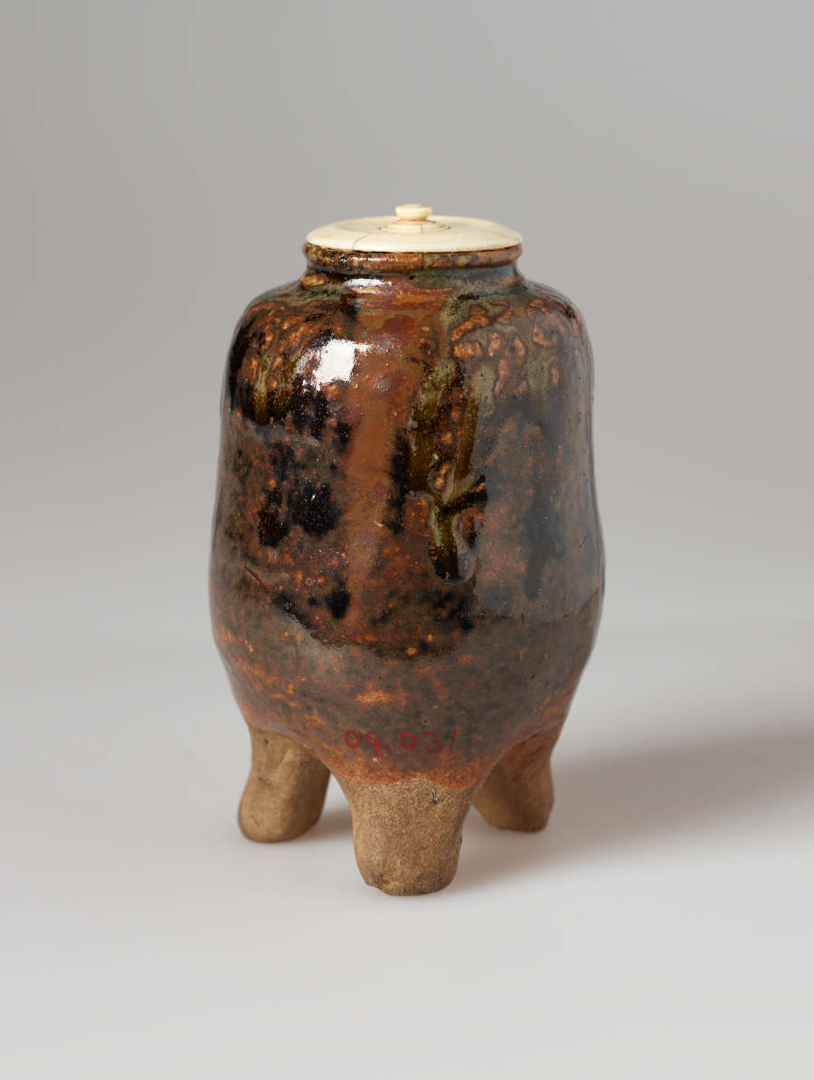 Small vessel in shades of reddish brown and black, standing on three rounded feet, with a cream colored lid.
