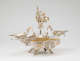 Elaborate silver double-oval-tray centerpiece with central winged-figurine holding up a gold garland, surrounded by cherubs, and framed by swirling arms extending to two shallow dishes, all elevated on four legs.