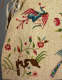 Detail of a cream colored fabric decorated by expressive embroidered motifs depicting a blue and red bird above red flowering buds growing out of earth with delicate stems and leaves.