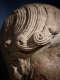 Close-up of a carved stone head focusing on curled locks of hair that coil at the end. The deep grooves are illuminated to highlight the craftsmanship. 