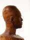 Close-up, three quarter back view of a textured, but glossy, wooden-carved bust depicting a male figure. The sculpture is dramatically lit, casting the face in shadow, while still retaining detail.