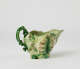  A short boat shaped pitcher green, cream, and brown in color with a sculptural handle and floral decorations.
