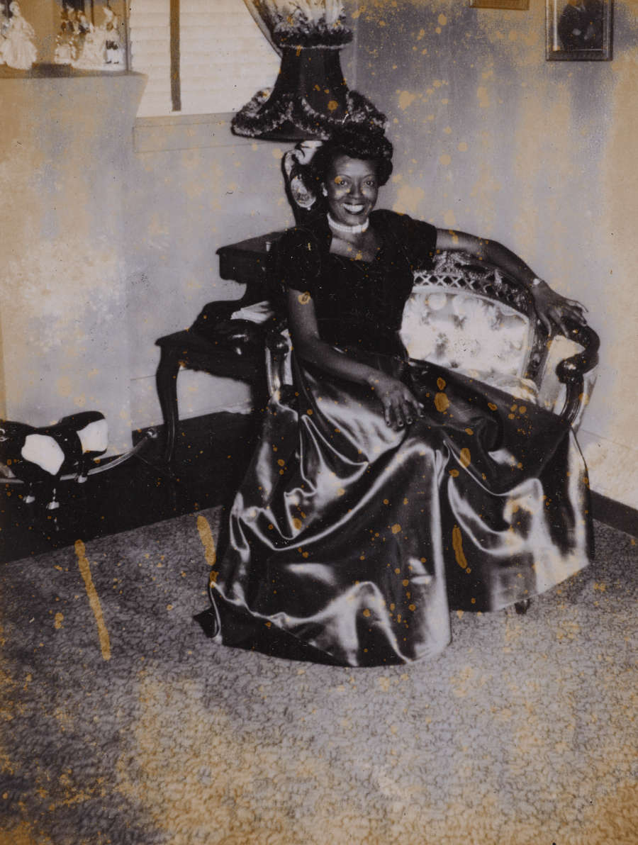 Photo of a dark-skinned woman wearing a satin gown. She in relaxing in an ornate armchair in a living room, smiling warmly at the camera. 