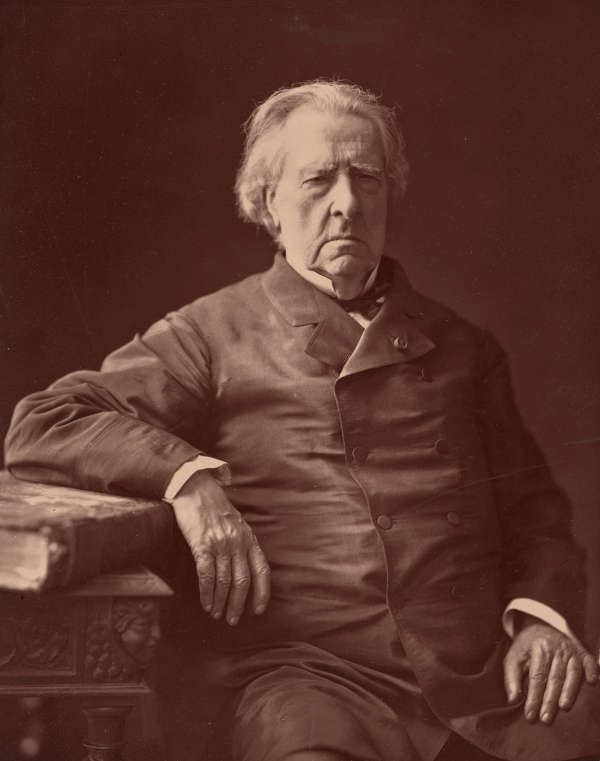 A warm, formal, black and white portrait of an older man seated gazing sternly at the viewer, wearing a buttoned coat with his right arm resting on a decorative table.