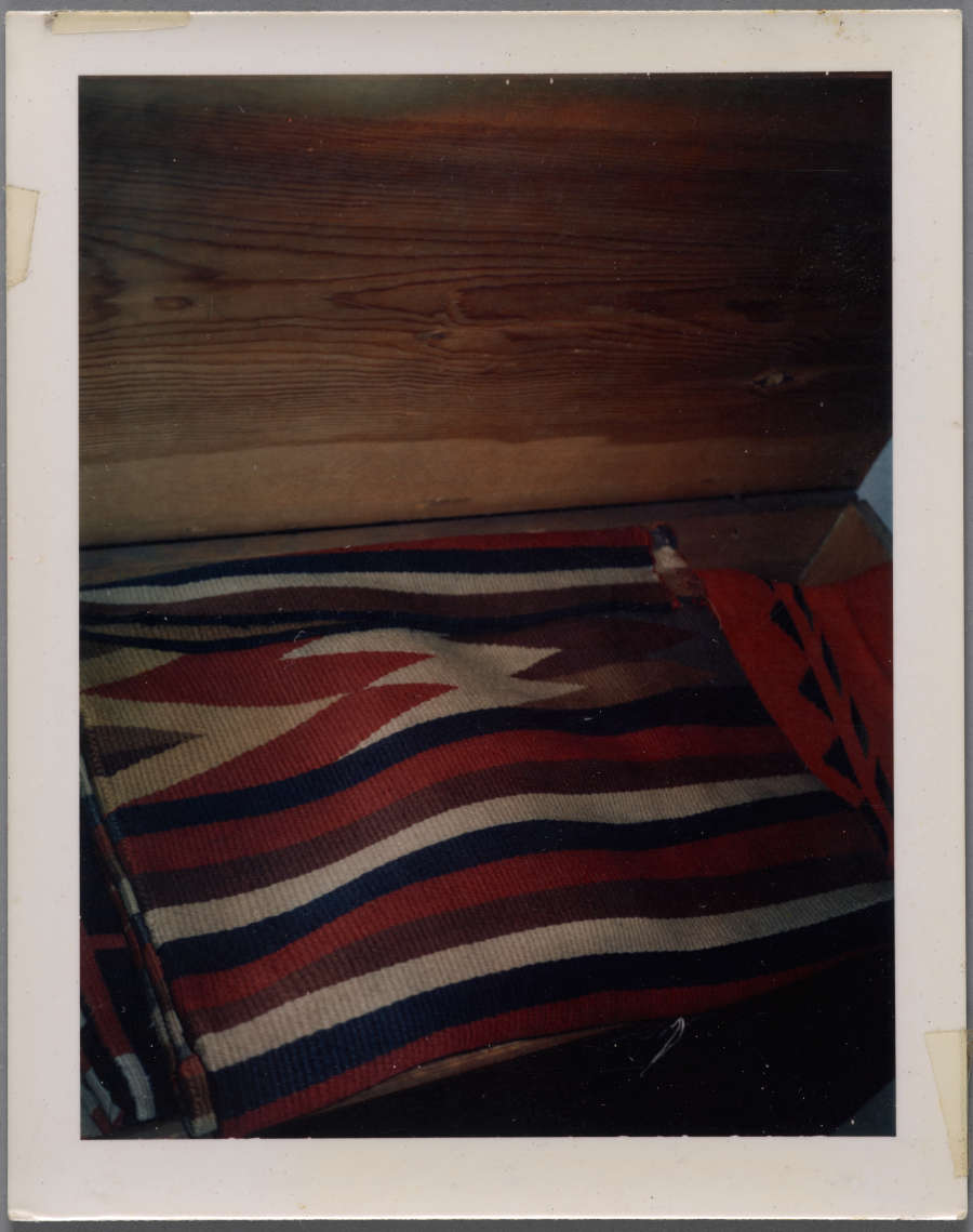 Vintage photograph showing a corner view of two blankets, a patterned red, blue, and beige blanket and a red woven blanket with green triangular designs, inside an open wooden box. 