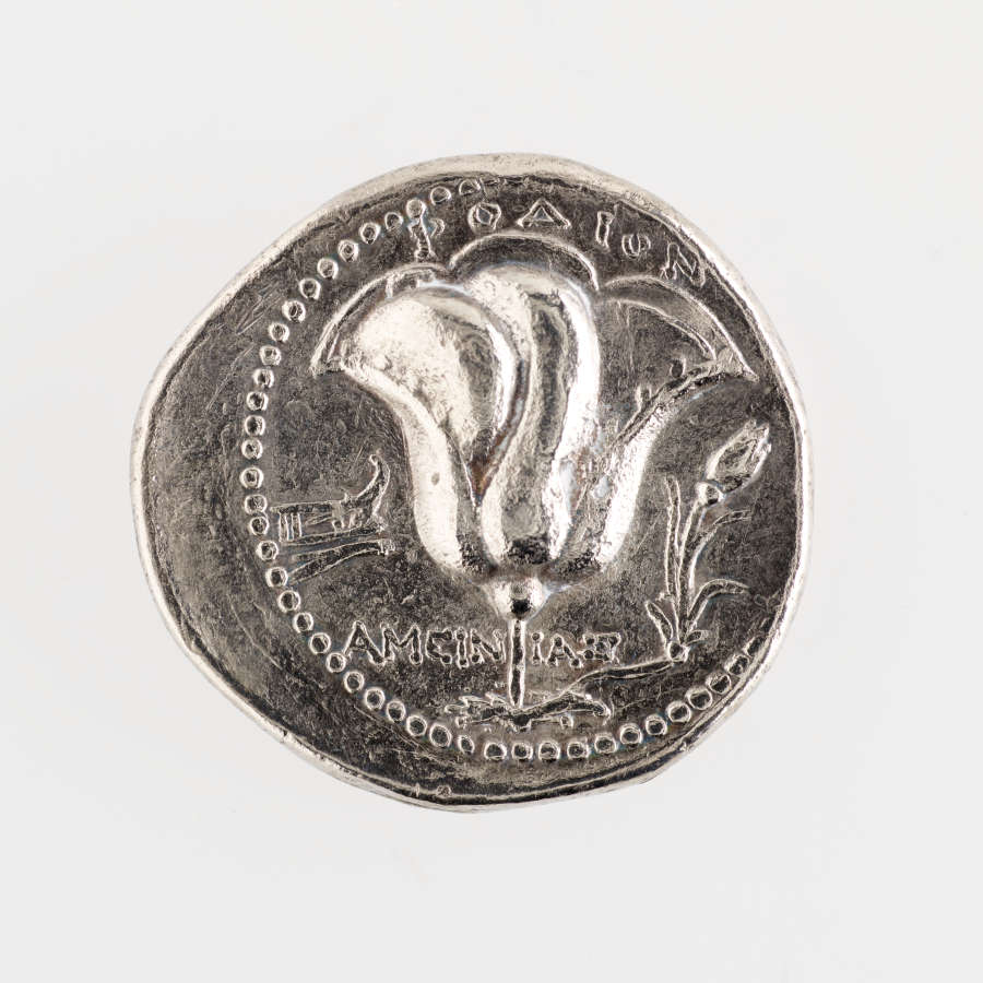Silver coin with irregular edges, embossed on its surface are small circles and a tulip as well as Greek lettering.