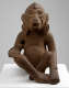Front of a brown sculpture monkey wearing earrings. Its tail drapes over the left arm, and its right arm holds its left foot. Its eyes and mouth are open.