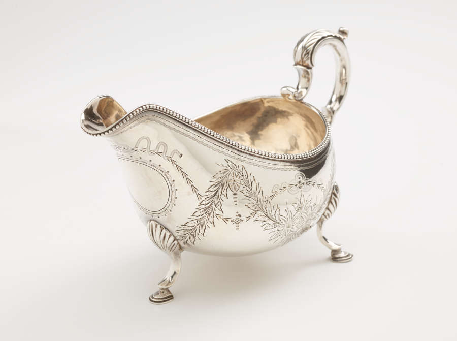 A silver sauce boat with a sculptural handle, high lip for pouring, and three protruding feet. There are floral engravings along the body of the vessel.