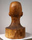 Back view of a carved wooden bust depicting the head of a male figure. The wood is cracked while retaining shine, the sculpture also features carving marks throughout.