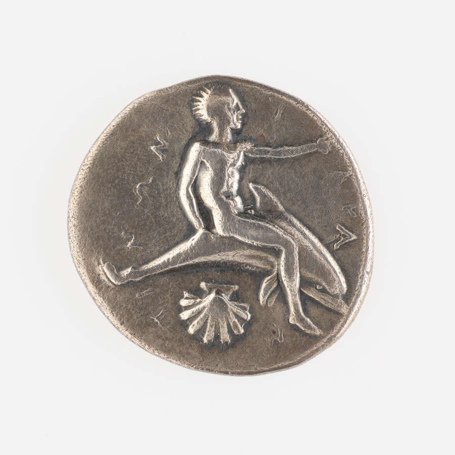 One side of a silver coin, embossed with an image of a nude man sitting on a dolphin, reaching to the right, above a clamshell. Lettering surrounds his figure.