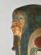 Side profile of a gilded coffin face with blue-outlined eyes, a sculpted false beard, and an ornate headdress with gold detailing and visible signs of age and wear.