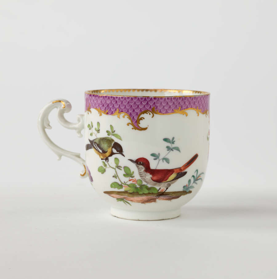 A white teacup with a swirling handle. Gold and pink decoration at top edge. Central design features two birds on a branch.