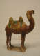 Side view of a glossy green, tan, and brown figurine of a camel with a long neck and two humps standing on a short attached rectangular platform.