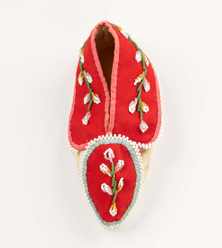 Moccasin top view. Wrapped round the heel is a red piece of fabric. The cap of the shoe features another ovular red fabric. Both feature white and green floral embroidery.